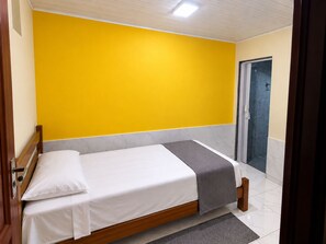 Una cama individual con estructura de madera, ropa de cama blanca y una manta gris en una habitación con paredes amarillas y piso de baldosas.