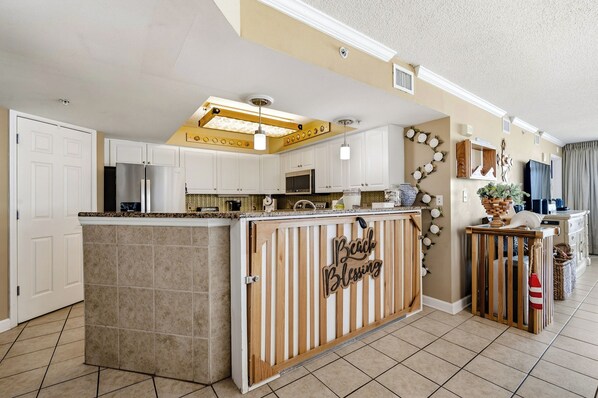 A kitchen with a central island, white cabinetry, and a decorative sign that reads 'Beach Blessing.' - Beach Blessing - Oceanfront Condo (North Myrtle Beach)
