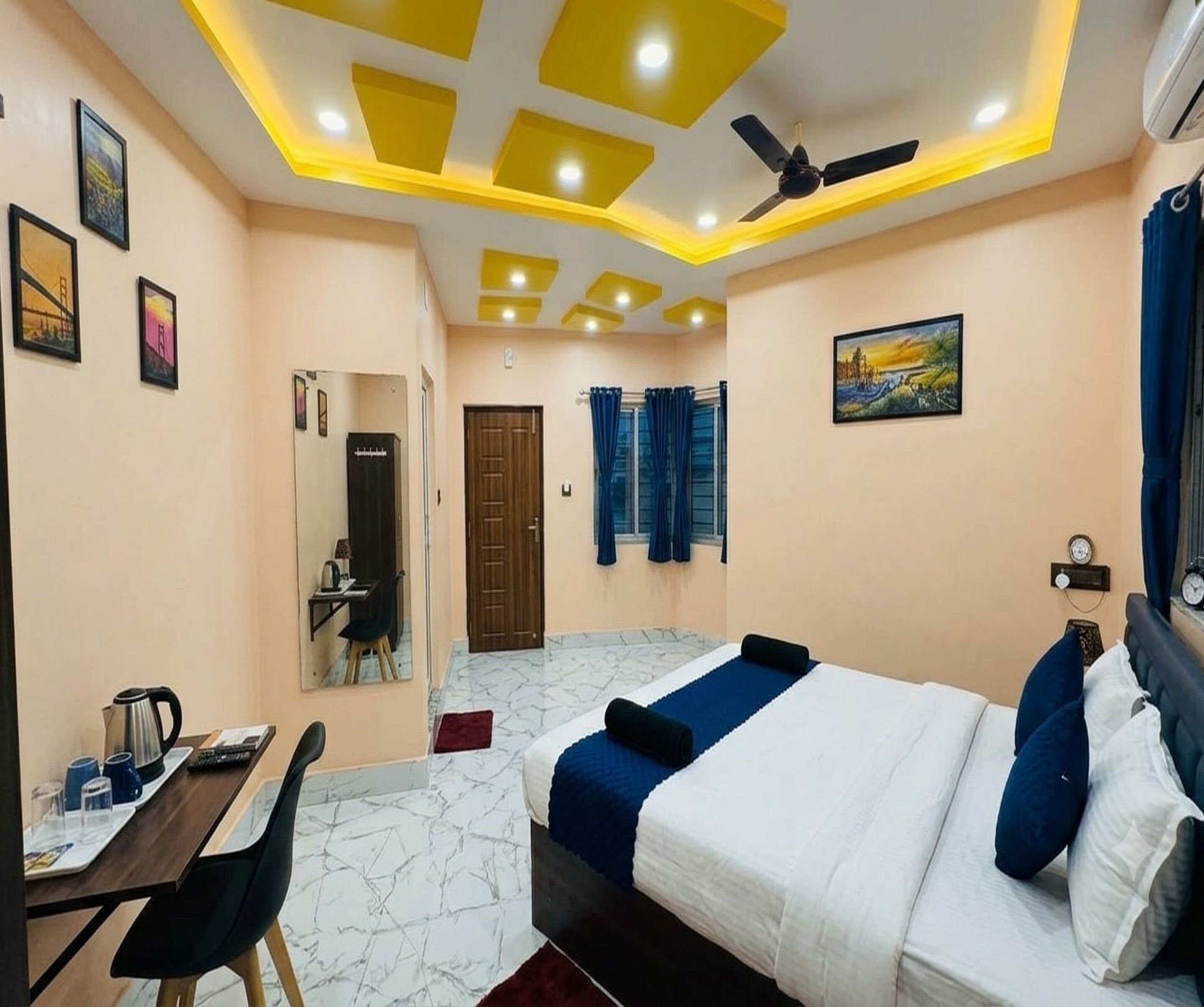 Habitación de hotel con cama, escritorio, silla, ventilador de techo y obras de arte en las paredes.