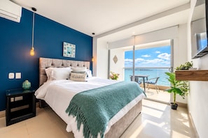 Quarto com cama, mesa de cabeceira, janela ampla com vista para o mar e varanda com mesa e cadeiras.