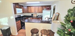 Cocina con armarios de madera, microondas, estufa, refrigerador y cafetera. Hay dos taburetes y un árbol de Navidad.