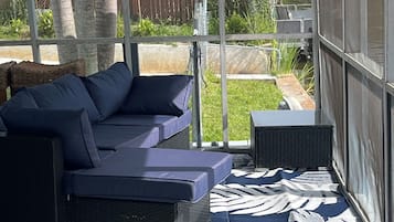 Un'area salotto esterna riparata, con un divano, poggiapiedi, un tappeto decorato e vista su un giardino recintato con alberi.