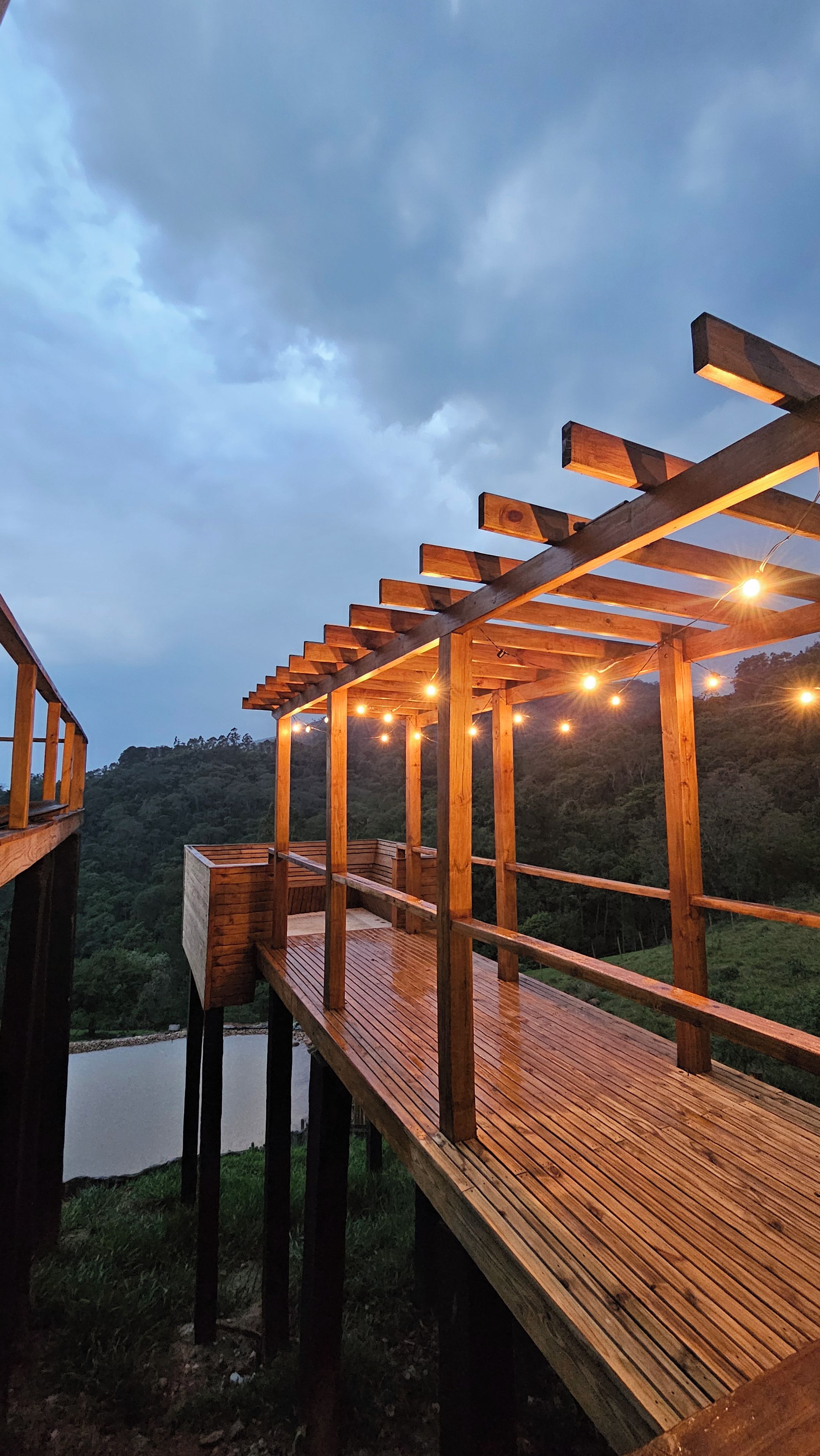 Um deck de madeira com estrutura de pérgola, cercado por paisagem natural.