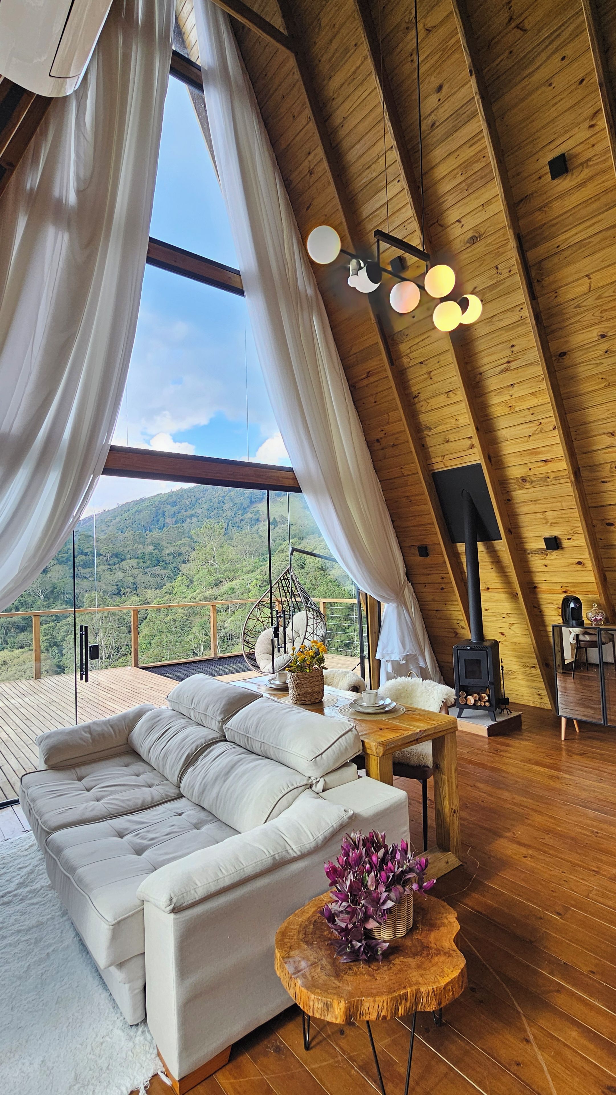 A cozy loft bedroom with a large window, a sofa, a wooden table, and a hanging chair.