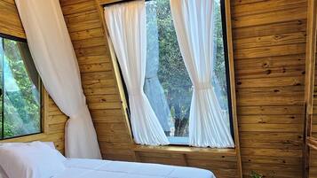 A bedroom with a wooden ceiling, a bed with white bedding, a window with curtains, and a plant on the floor.