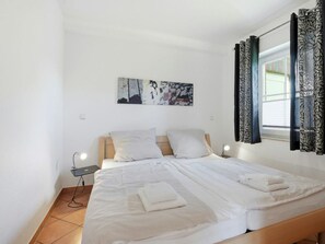 Ein Schlafzimmer mit Bett, Kissen, Nachttisch, Lampe, einem Fenster mit Vorhängen und einem Wandbild.