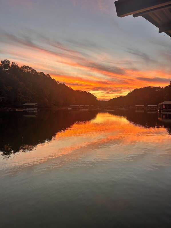 A calm river reflecting a sunset sky, surrounded by forested hills and small buildings. - New listing discount! Sunny Shores on Hartwell- Dock, kayaks, pedal boat, games (Martin)