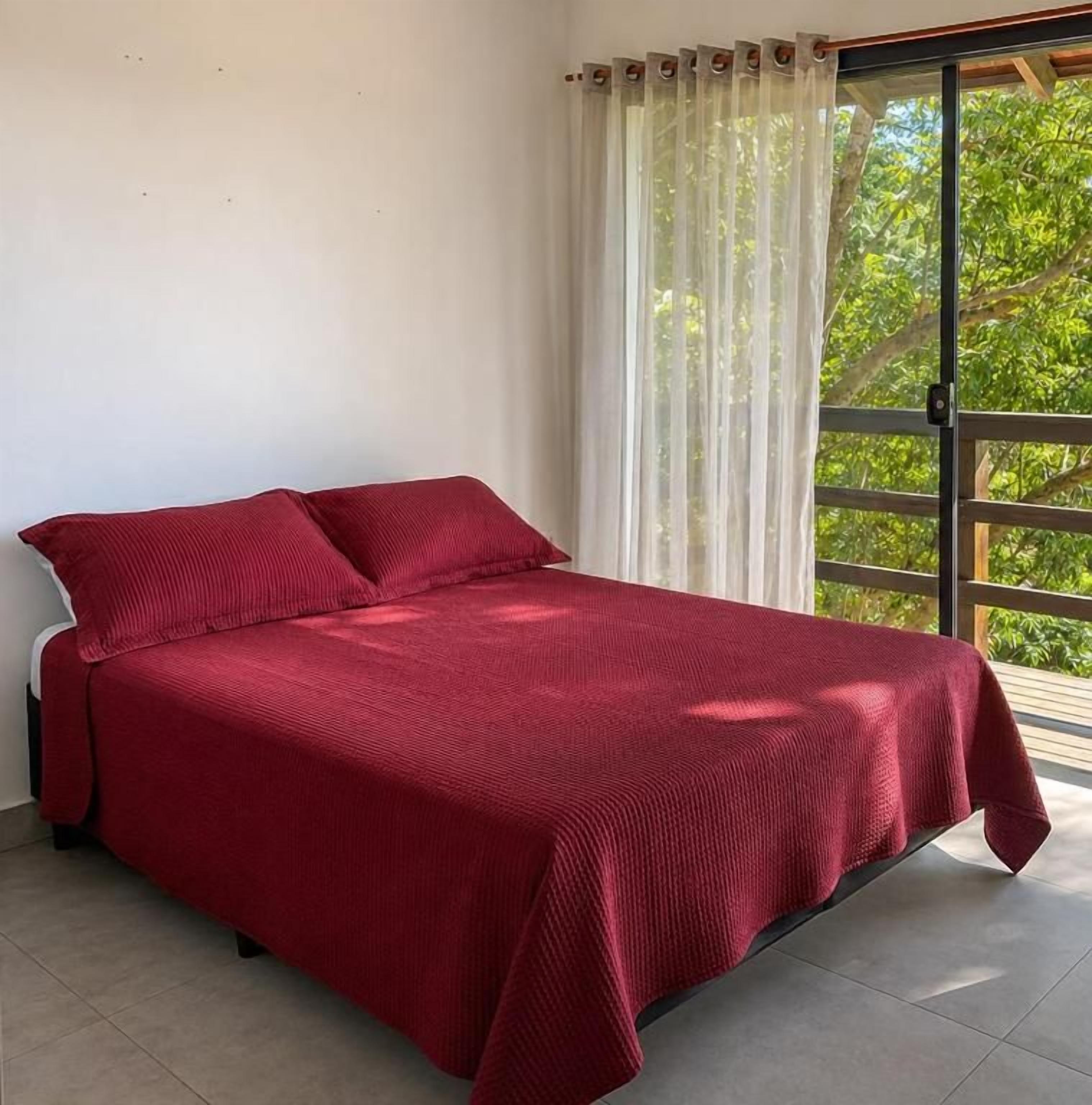 A bed with a red cover in a room with a large window and a view of greenery outside.