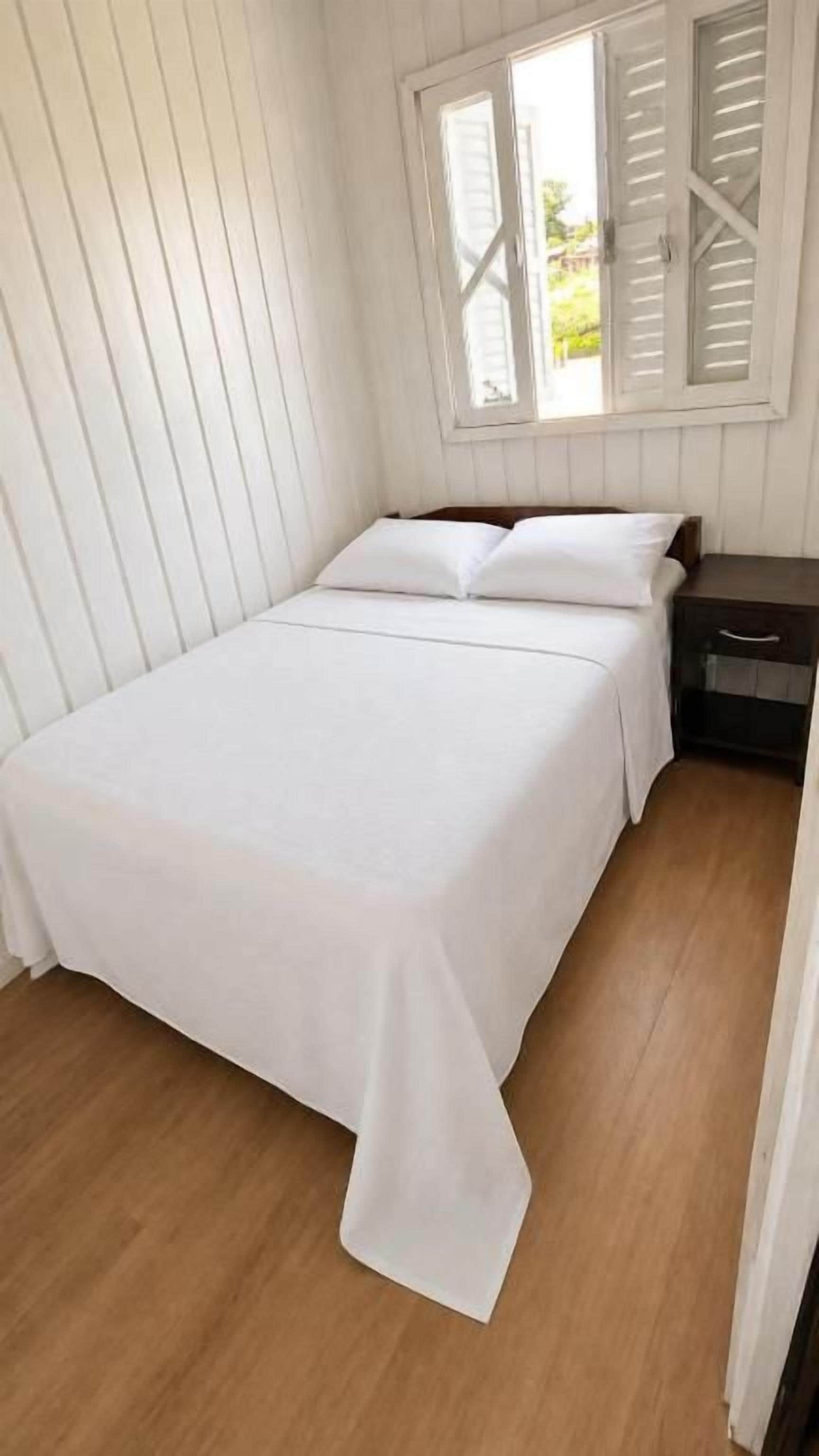 Una cama individual con una colcha blanca en una habitación con suelo de madera y paredes blancas.