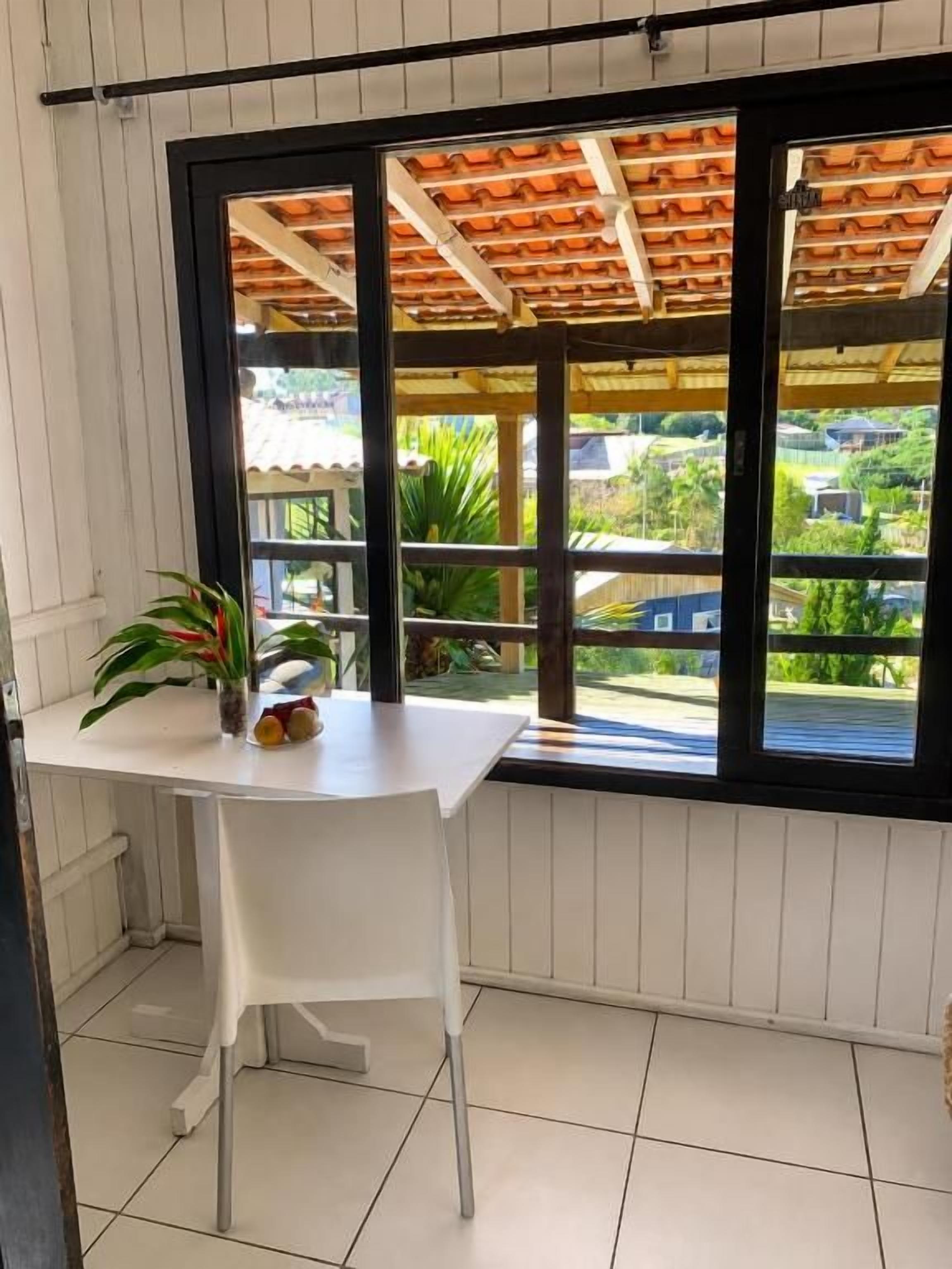 A balcony with a white table and chair, a potted plant, and a view of greenery and buildings.