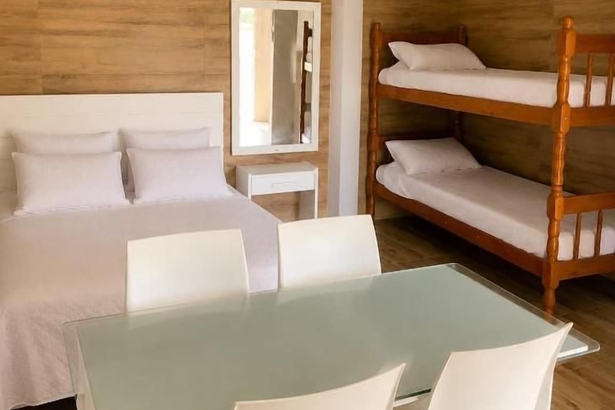A room with a bunk bed, a single bed, a glass-top table with chairs, a bedside table, and a mirror.