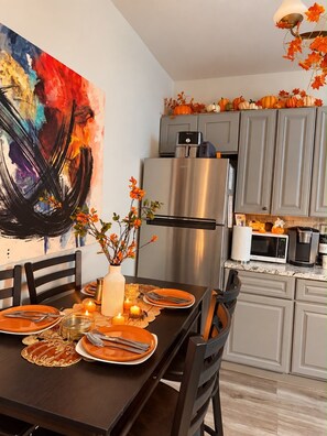 A modern kitchen with grey cabinets, a stainless steel refrigerator, and a dining table set for four with orange plates and candles.