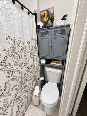 Ein Badezimmer mit Toilette, einer Duschkabine mit Blumenmuster, einem grauen Schrank mit Regalen und einem gerahmten Kunstwerk über der Toilette.