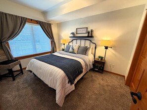 A bedroom with a bed, two bedside tables, a chair, a window with curtains, and a wall-mounted shelf with decorative items. - GB  102: Beautiful water view! Great Birch Bay location, free Wi-Fi. (Birch Bay)