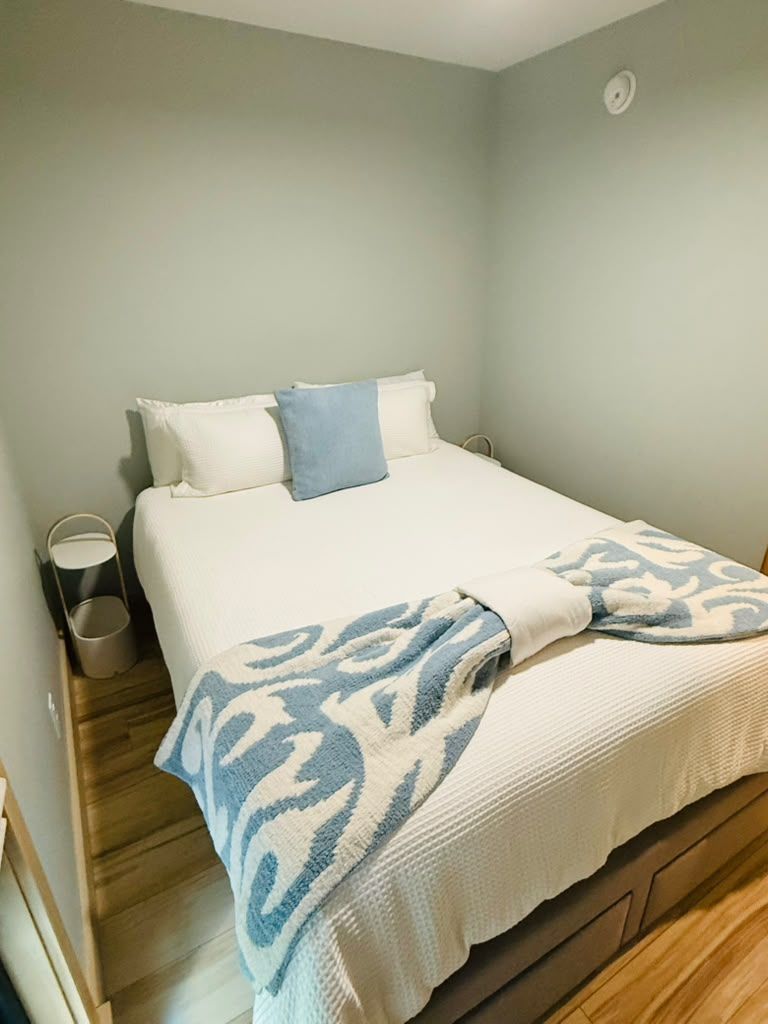 A single bed with a white and blue patterned blanket, a small bedside table with a lamp, and a wall-mounted light fixture.
