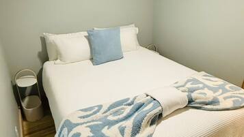 A single bed with a white and blue patterned blanket, a small bedside table with a lamp, and a wall-mounted light fixture.