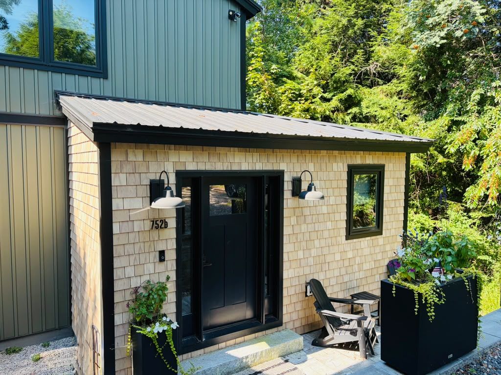 A small brick building with a black door and windows, a patio with a table and chairs, and a garden with plants.