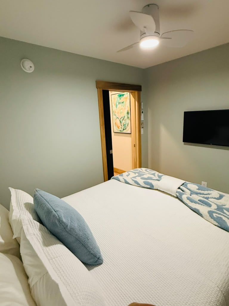 A bedroom with a bed, pillows, a ceiling fan, a wall-mounted TV, and a door leading to another room.
