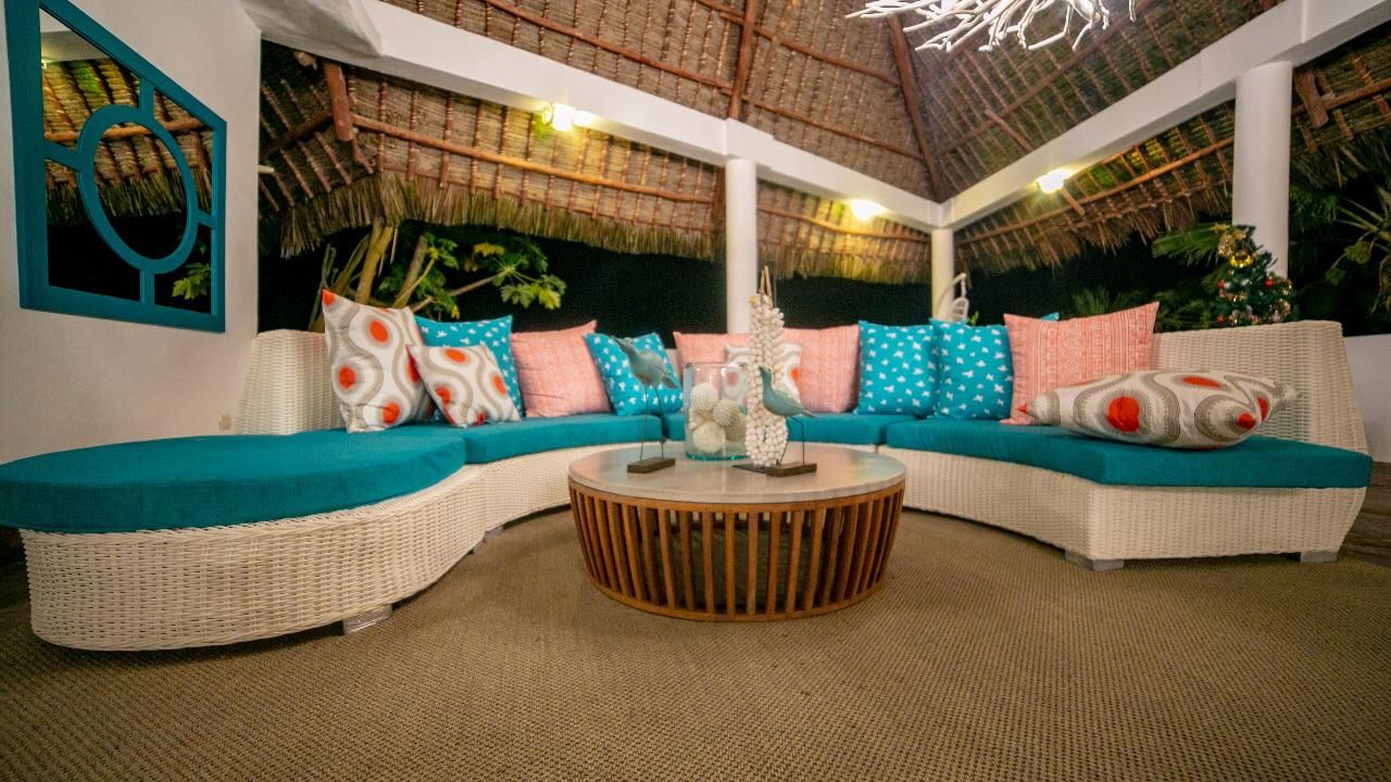 Ein gebogenes, weißes Wicker-Sofa mit türkisfarbenen Kissen und gemusterten Zierkissen, ein Couchtisch aus Holz und eine mit Stroh gedeckte Decke.