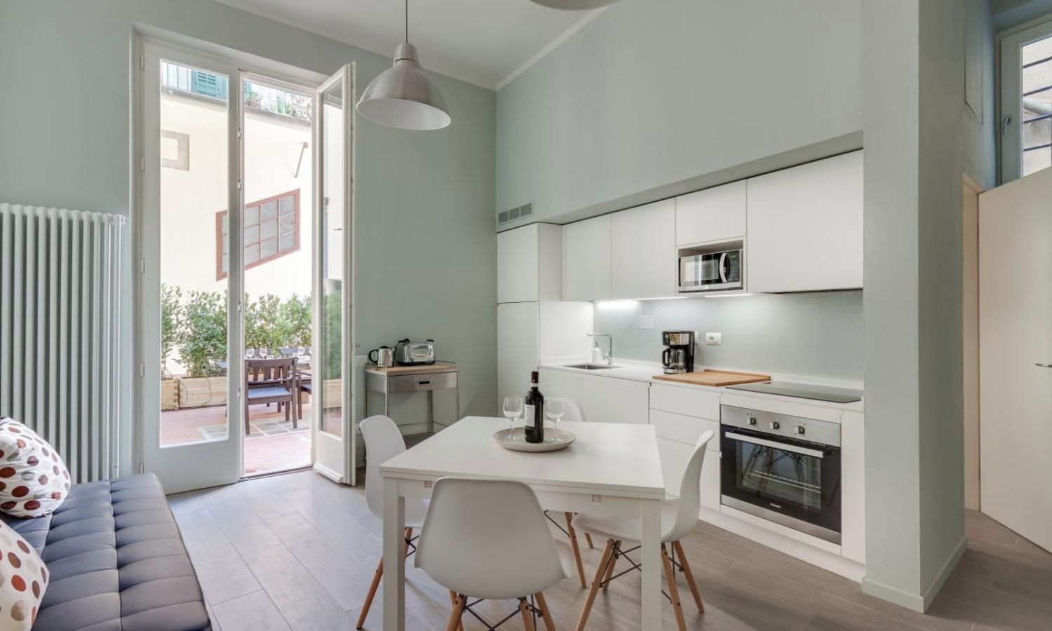 A modern kitchen with white cabinets, a microwave, and an oven. There is a dining area with a table and chairs, and a sofa with a polka dot cushion.