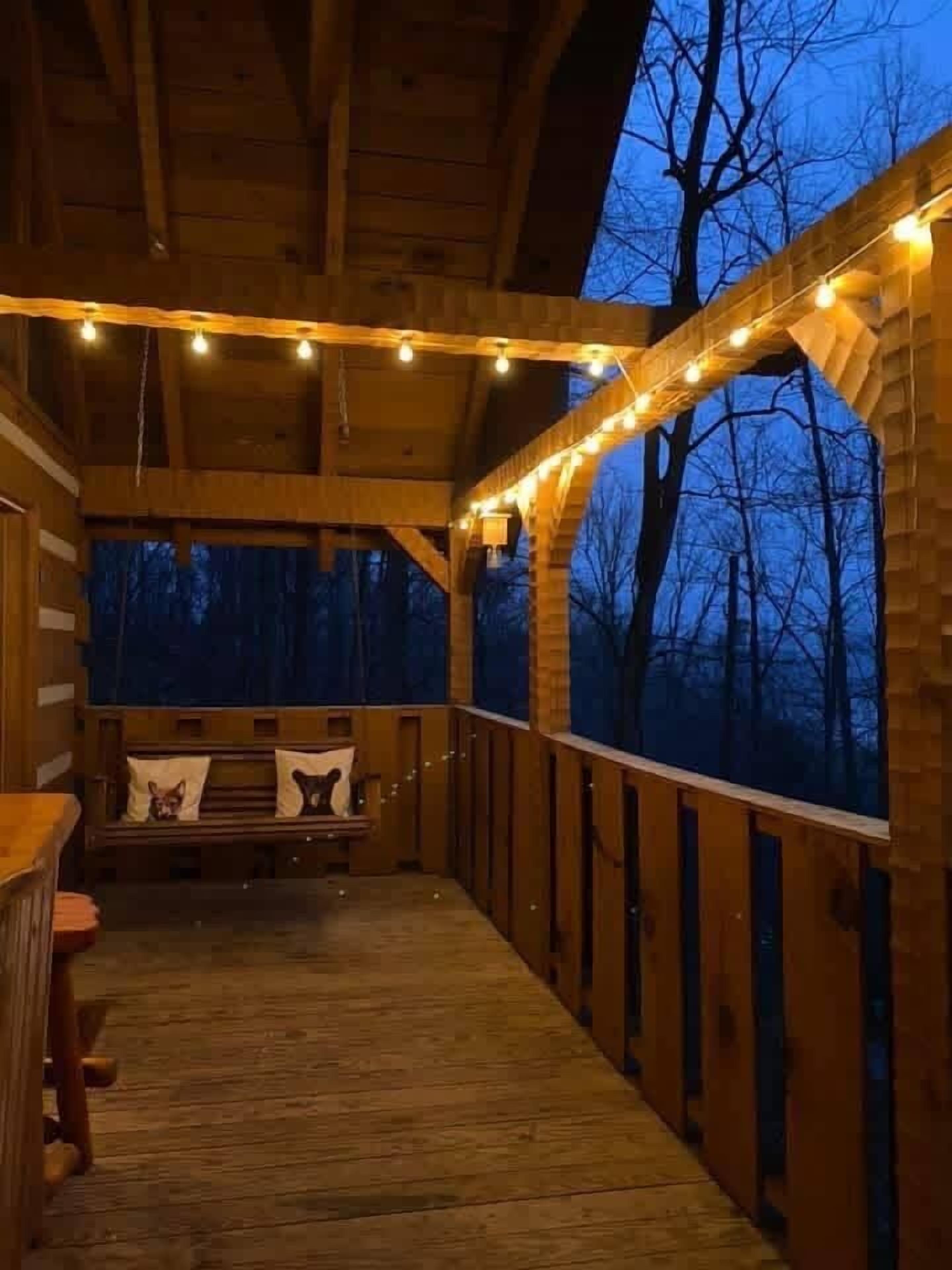 A wooden deck with string lights, a bench with cushions, and a view of trees.