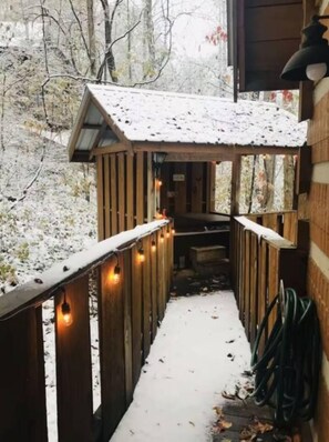 Una terraza de madera con un sendero cubierto de nieve que lleva a un edificio, rodeado de árboles.