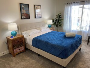 Quarto com cama, duas mesas de cabeceira com abajures, uma planta em vaso e duas imagens emolduradas na parede.