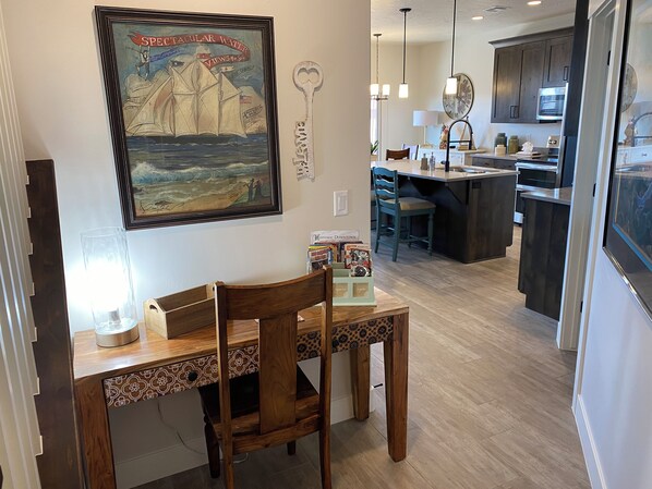 Uma área de refeições com uma mesa e cadeiras de madeira, um quadro na parede e uma cozinha ao fundo.