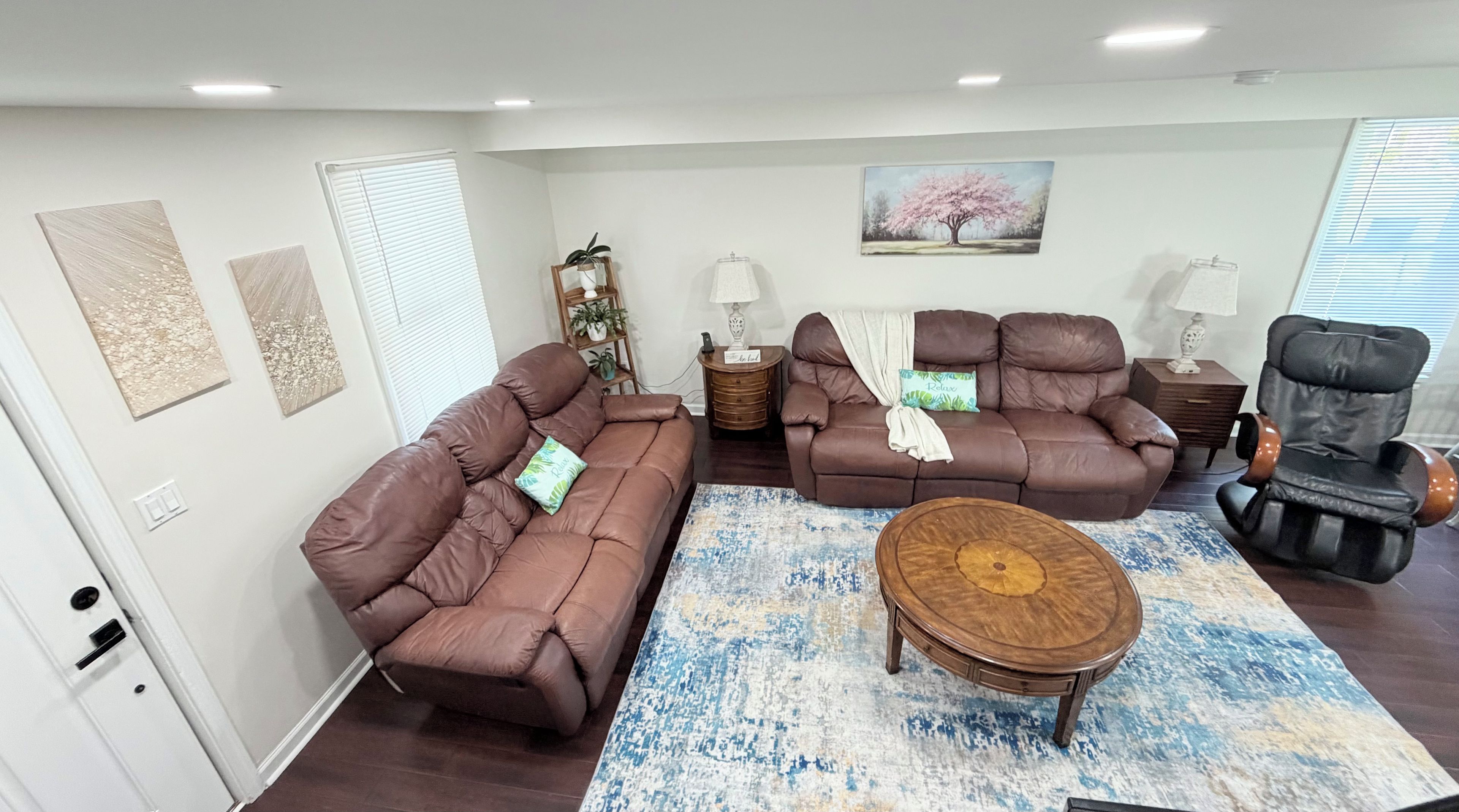 Ein Wohnzimmer mit zwei braunen Sofas, einem Holztisch und einem Massagesessel. An den Wänden befinden sich Kunstwerke und ein Baumgemälde.