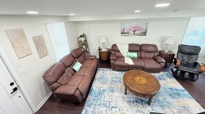 A living room with two brown sofas, a wooden coffee table, and a massage chair. There are wall art pieces and a painting of a tree.