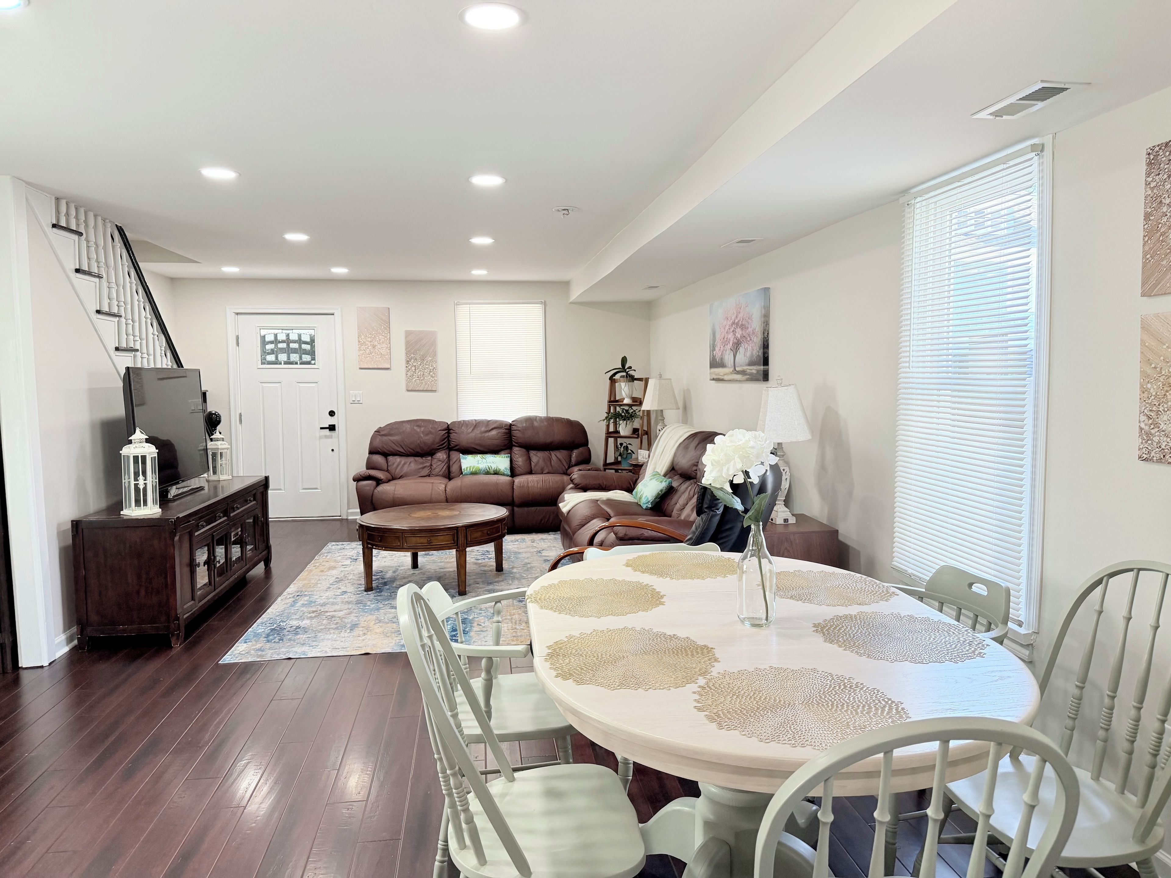 A spacious living room with a brown leather sofa, a dining table with white chairs, and a staircase leading to another level.