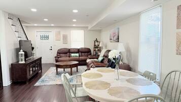 A spacious living room with a brown leather sofa, a dining table with white chairs, and a staircase leading to another level.