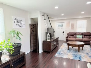 A living room with a brown leather sofa, a coffee table, a TV on a wooden cabinet, a potted plant, and a staircase leading to another room.