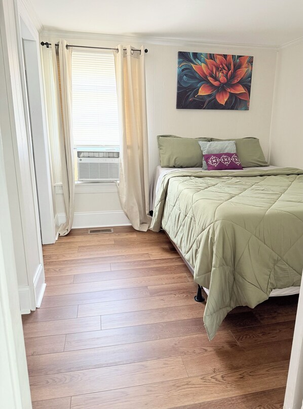 A bedroom with a bed, a window with curtains, a radiator, and a painting on the wall.
