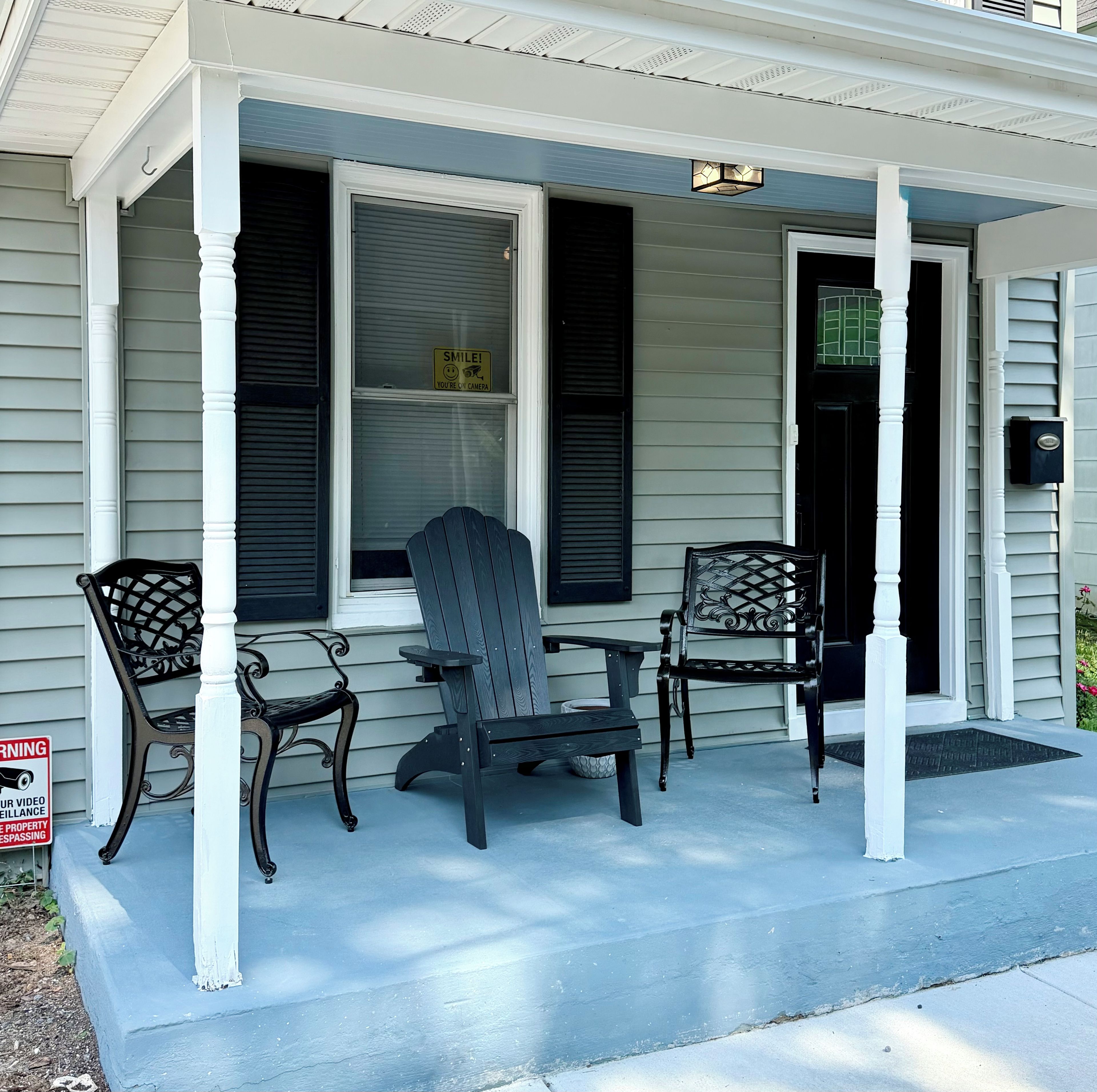 A porch with two Adirondack chairs and a table, a black door, and a window with a 'SMILE! YOU'RE ON CAMERA' sign.