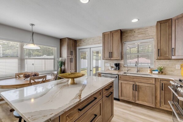 A modern kitchen with wooden cabinets, a marble countertop, and a dining area with a round table and chairs. - Hot Tub-Heated Pool & Fenced Yard for your Pup! (New Smyrna Beach)