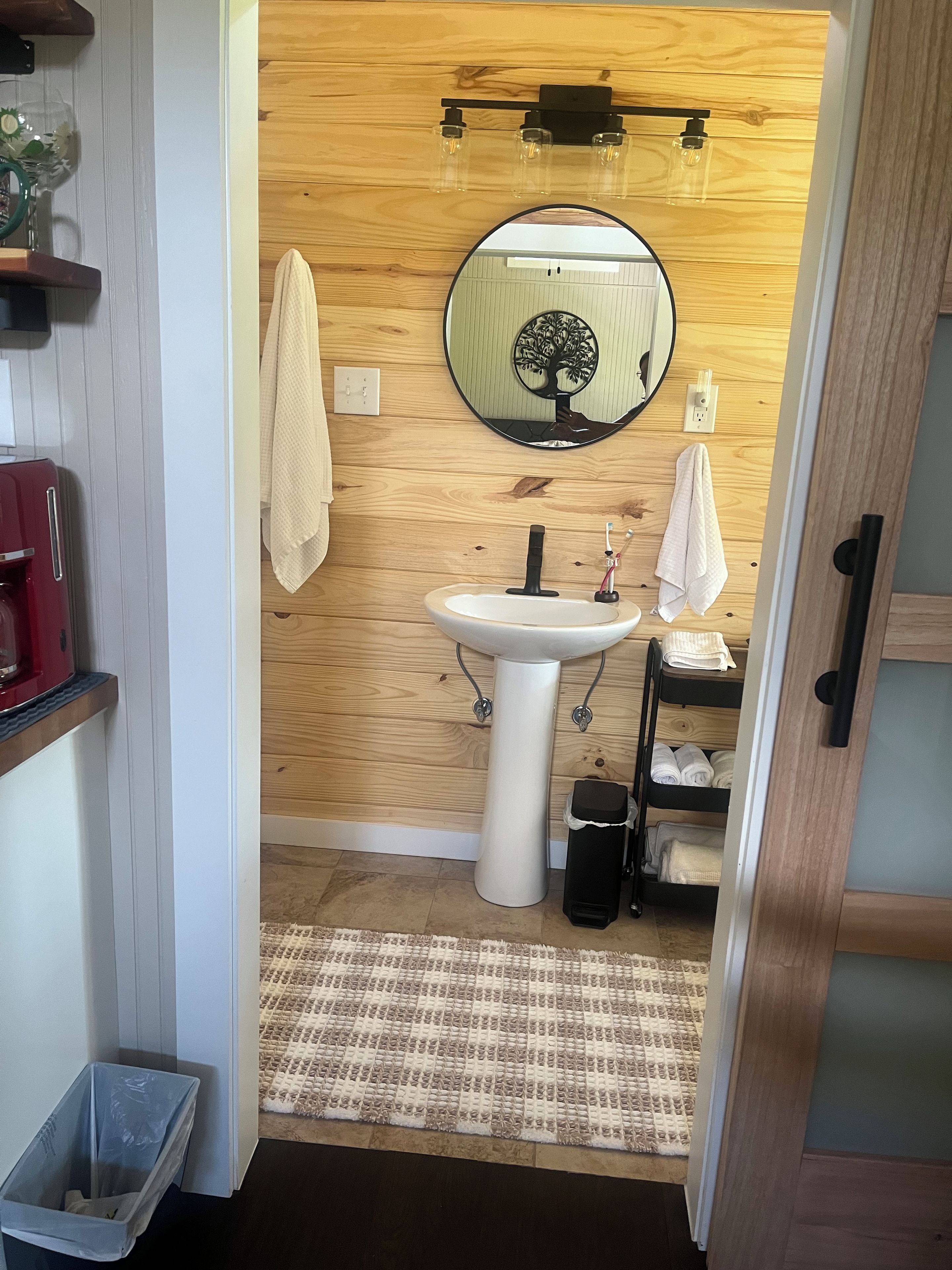 Een kleine badkamer met een witte wastafel, een ronde spiegel met een ontwerp van een boom, een handdoekhouder en een houten wand.