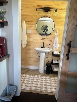 A compact bathroom with a white pedestal sink, a round mirror with a tree design, a towel rack, and a wooden wall.