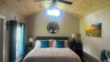 Un dormitorio con cama, ventilador de techo, adorno de árbol en la pared, mesita de noche con lámpara y un cuadro de un campo de girasoles.