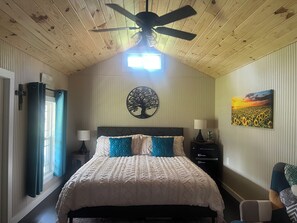 A bedroom with a bed, a ceiling fan, a wall-mounted tree decoration, a nightstand with a lamp, and a painting of a sunflower field. - Studio TINY HOUSE 3 blocks from Jack Daniel Distillery, 2 blocks to town square (Lynchburg)