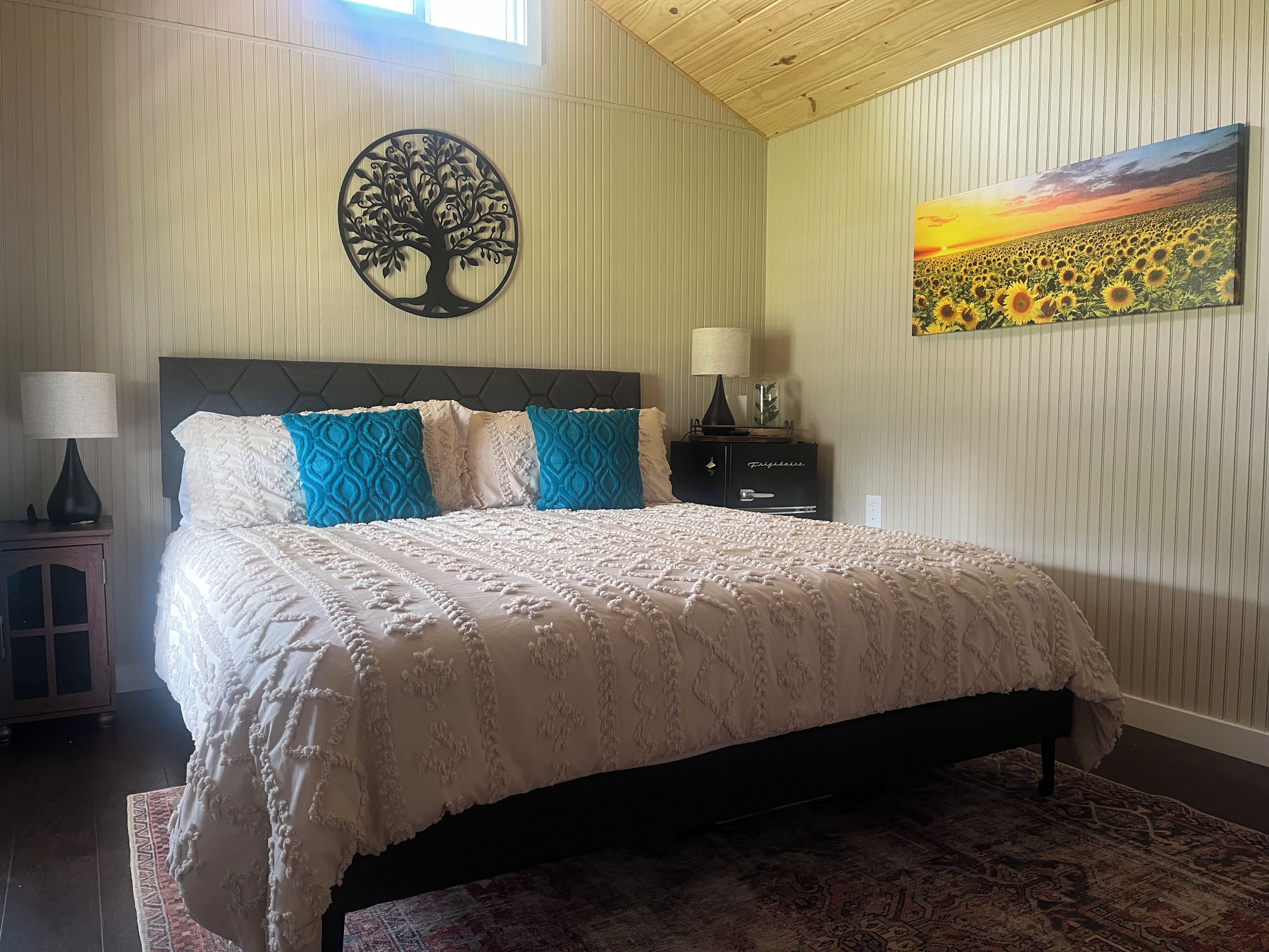 A bedroom with a bed, a nightstand, a lamp, a dresser, a wall art piece, and a rug.