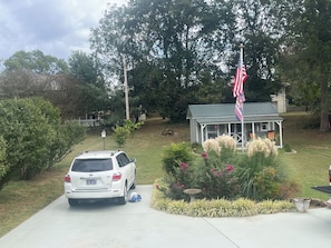 Un camino de entrada residencial con una camioneta SUV blanca estacionada, una casa pequeña con porche y un asta con la bandera estadounidense.
