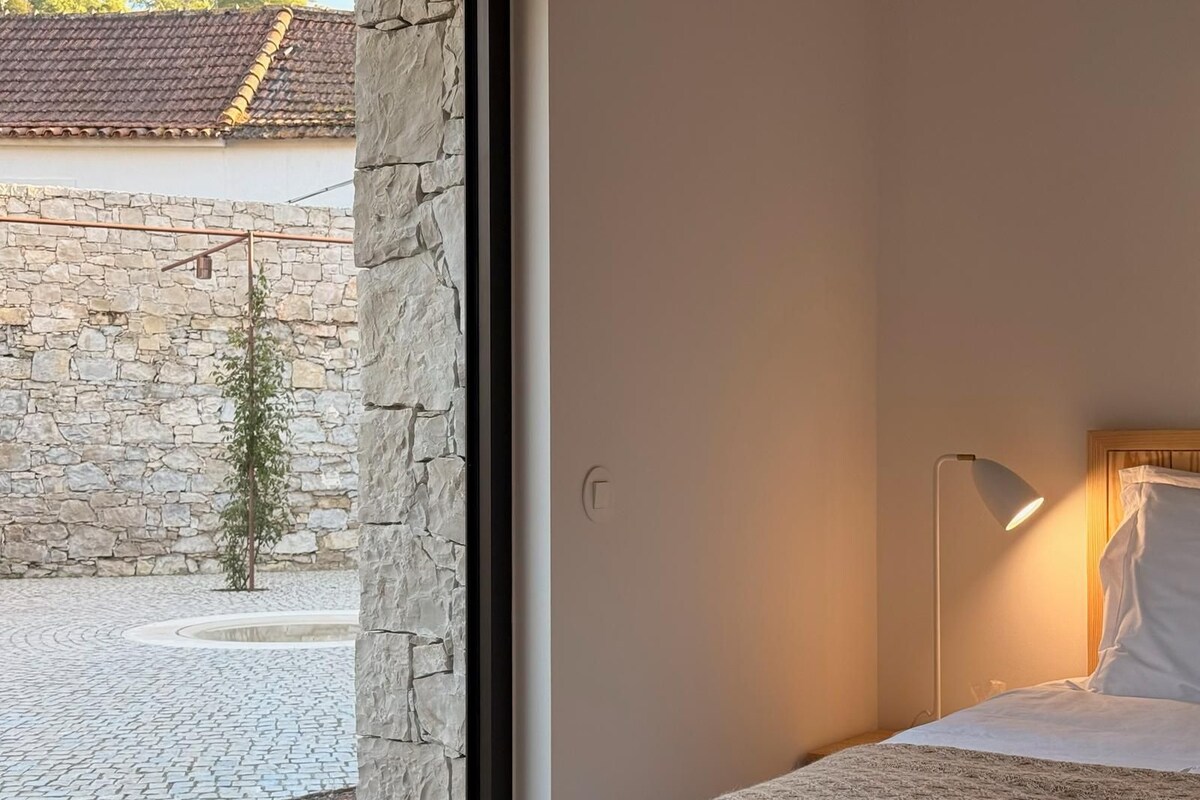 Ein Schlafzimmer mit einem Bett, einer Nachttischlampe und Blick auf einen Innenhof mit einer Steinmauer und einem Baum.