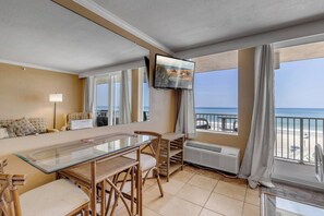 Ein Hotelzimmer mit Balkon, einem an der Wand montierten Fernseher, einem Glastisch mit Stühlen und Blick auf den Strand.