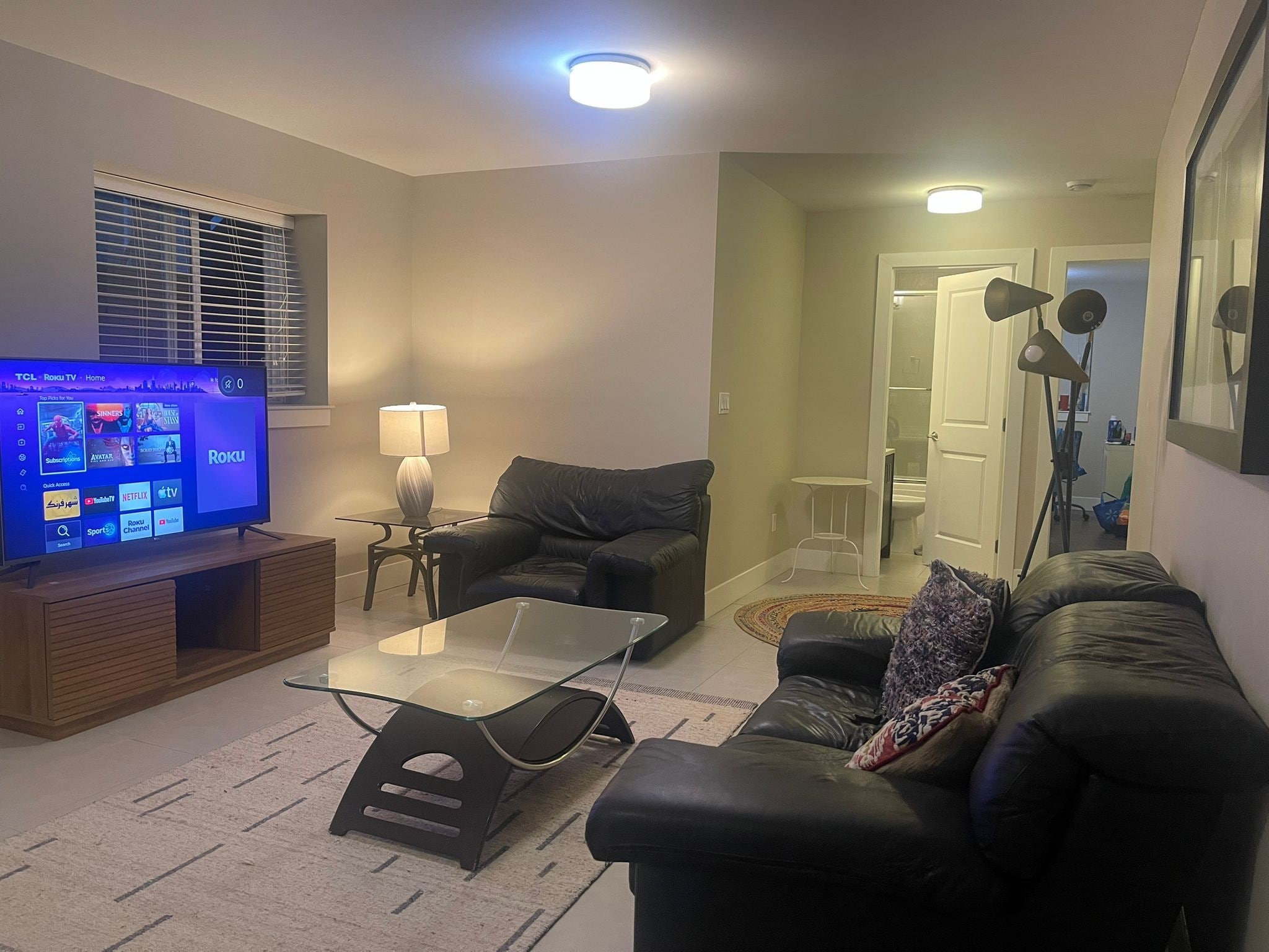 En stue med et fladskærms-tv, et glasbord, to sorte lædersofaer, en lampe og en dør til et andet rum.