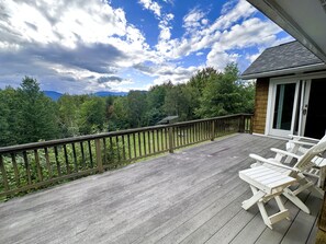 A wooden deck with white chairs, a railing, and a view of a forested area. - Valeside Lodge: Whiteface views, firepit, privacy, woodstove, rec room (Jay)