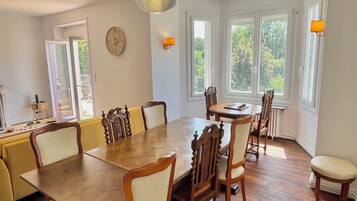 A dining area with a wooden table and chairs, a chandelier, and large windows with a view of trees.