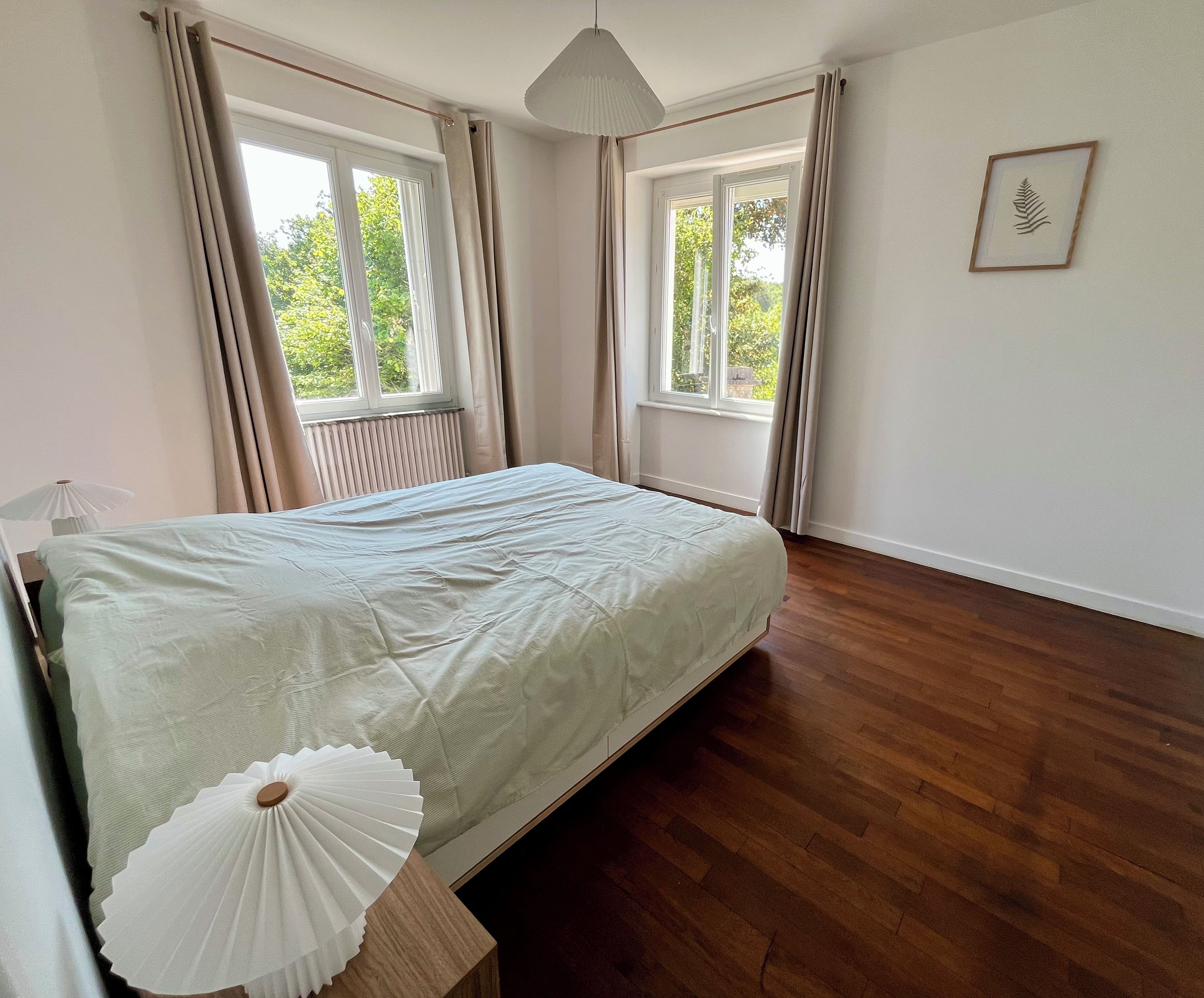 A bedroom with a bed, wooden flooring, and two windows with curtains.