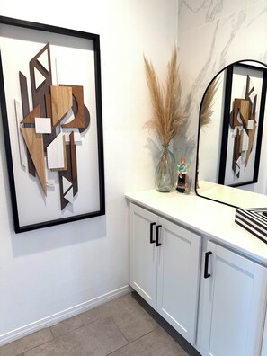 A bathroom with a framed abstract art piece, a vase with pampas grass, and a mirror with a geometric design. - La Quinta Oasis w/Pool, Hot Tub, Outdoor Kitchen, 4bedrooms/4baths (La Quinta)