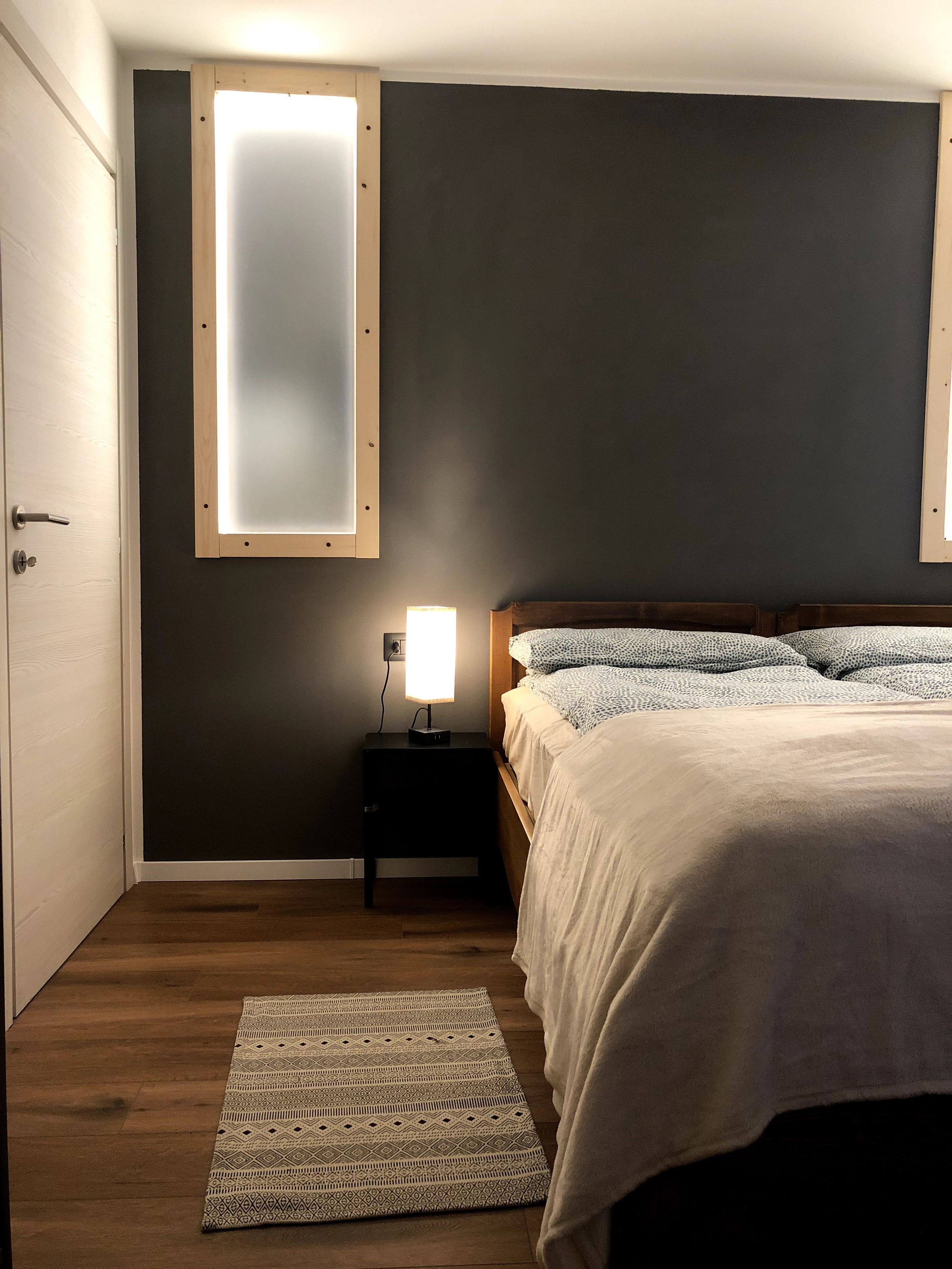 Ein Schlafzimmer mit einem Bett, einem Nachttisch mit Lampe, einem Fenster mit Jalousien und einem Holzboden.